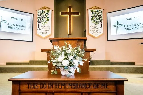 The sanctuary at Arbor Heights Bible Church: the pulpit and a wooden cross, with the communion table reading &ldquo;This do in remembrance of me&rdquo;